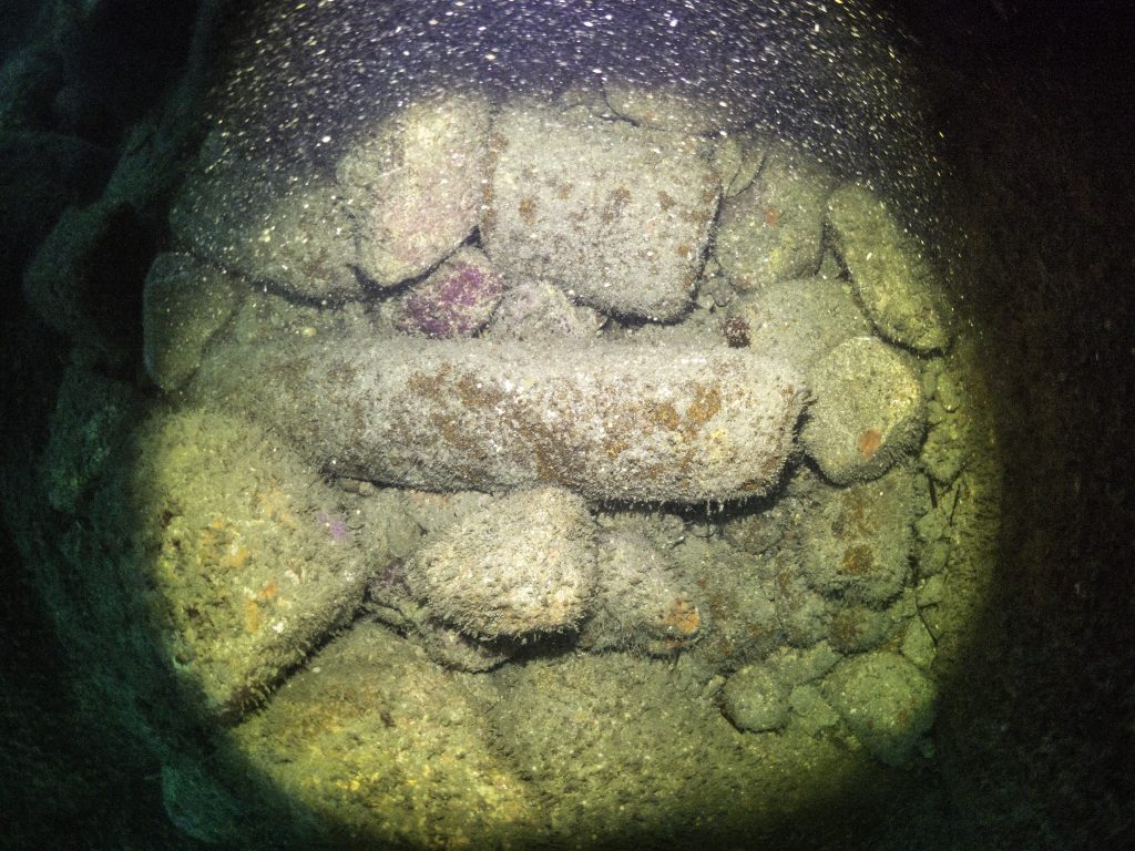 Cannon sitting on a rock strewn seabed inside a small cave formed by large boulders. This image is illuminated by torch light