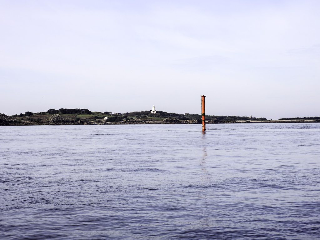 Metal pole rising from the sea surface. The island of St Agnes is visible in the background where the white painted lighthouse is visible