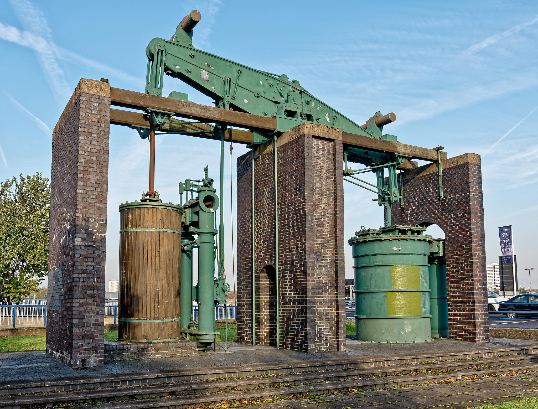 Photograph of the Grazebrook blowing engine built in 1817 against a blue sky. A red brick structure of three supporting walls supports a green iron beam on the central wall. On either side of the central wall is a large cylinder. The left hand cylinder is cased in wood, and is 42 inches in diameter. The Right hand cylinder is painted green and is an 84 inch air pump.