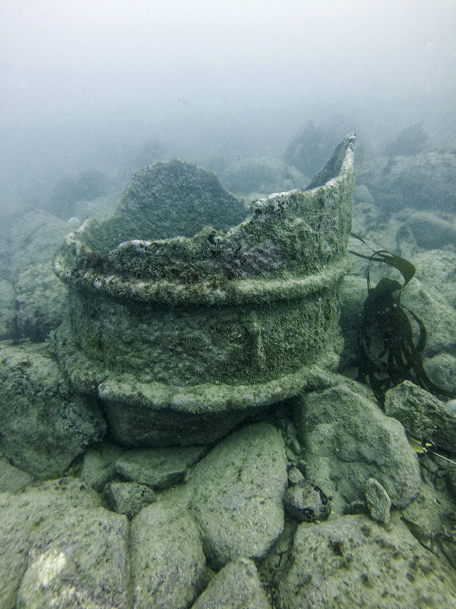 An underwater photograph of cylinder fragment C6 lying upright on the rocky seabed. The flange at the bottom and one of the reinforcing rings can be seen clearly.